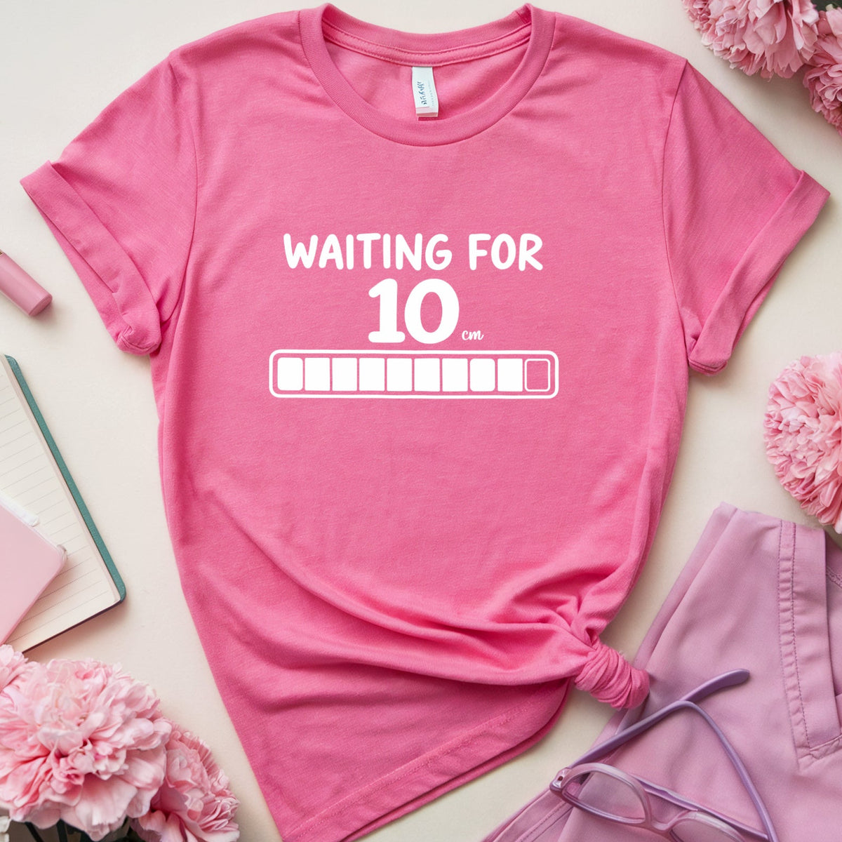 A pink graphic T-shirt laid flat on a light beige surface, surrounded by pink and white flowers, a notebook, pen, lipstick, and lavender scrubs with glasses. The shirt has white text reading 'WAITING FOR 10.