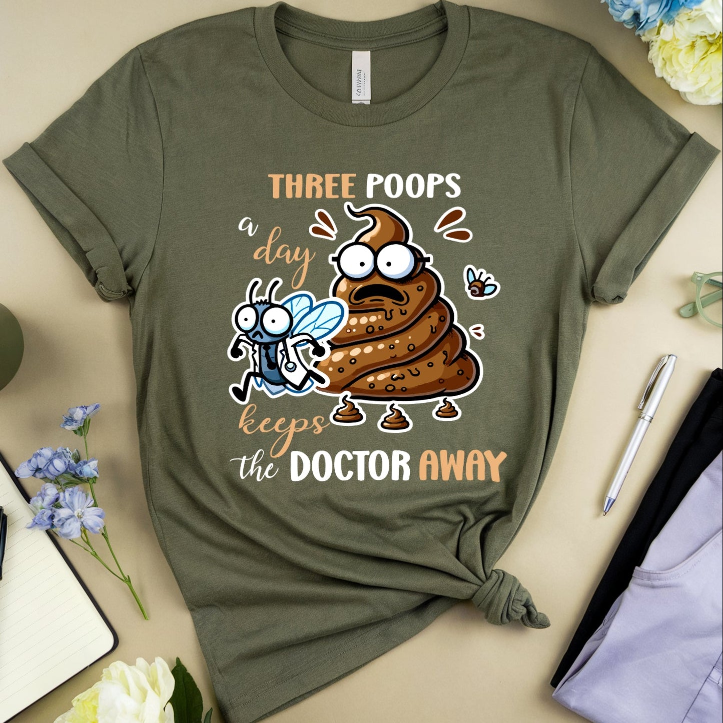 Green t-shirt with cartoon poop emoji and text on a beige surface with flowers and a mug.