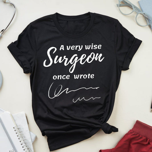 Black t-shirt for surgeons with 'A very wise surgeon once wrote' text on a light surface with medical-themed items.