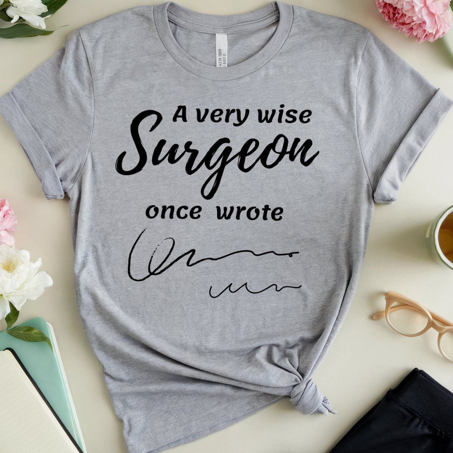Gray t-shirt for surgeons with humorous text on a table with flowers and a cup.