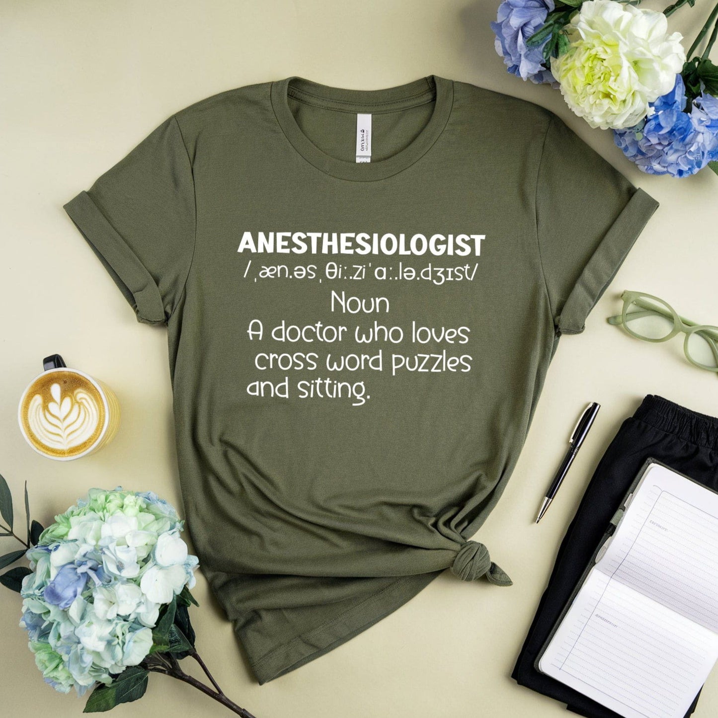 Green t-shirt with 'ANESTHESIOLOGIST' definition text on a light surface with flowers and a cup.