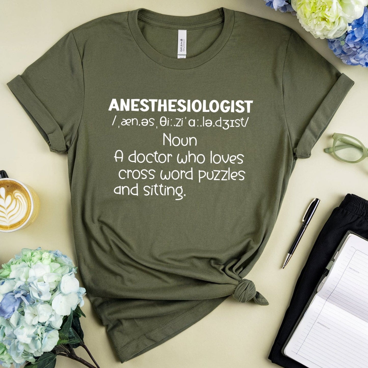 Green t-shirt with 'ANESTHESIOLOGIST' definition text on a light surface with flowers and a cup.