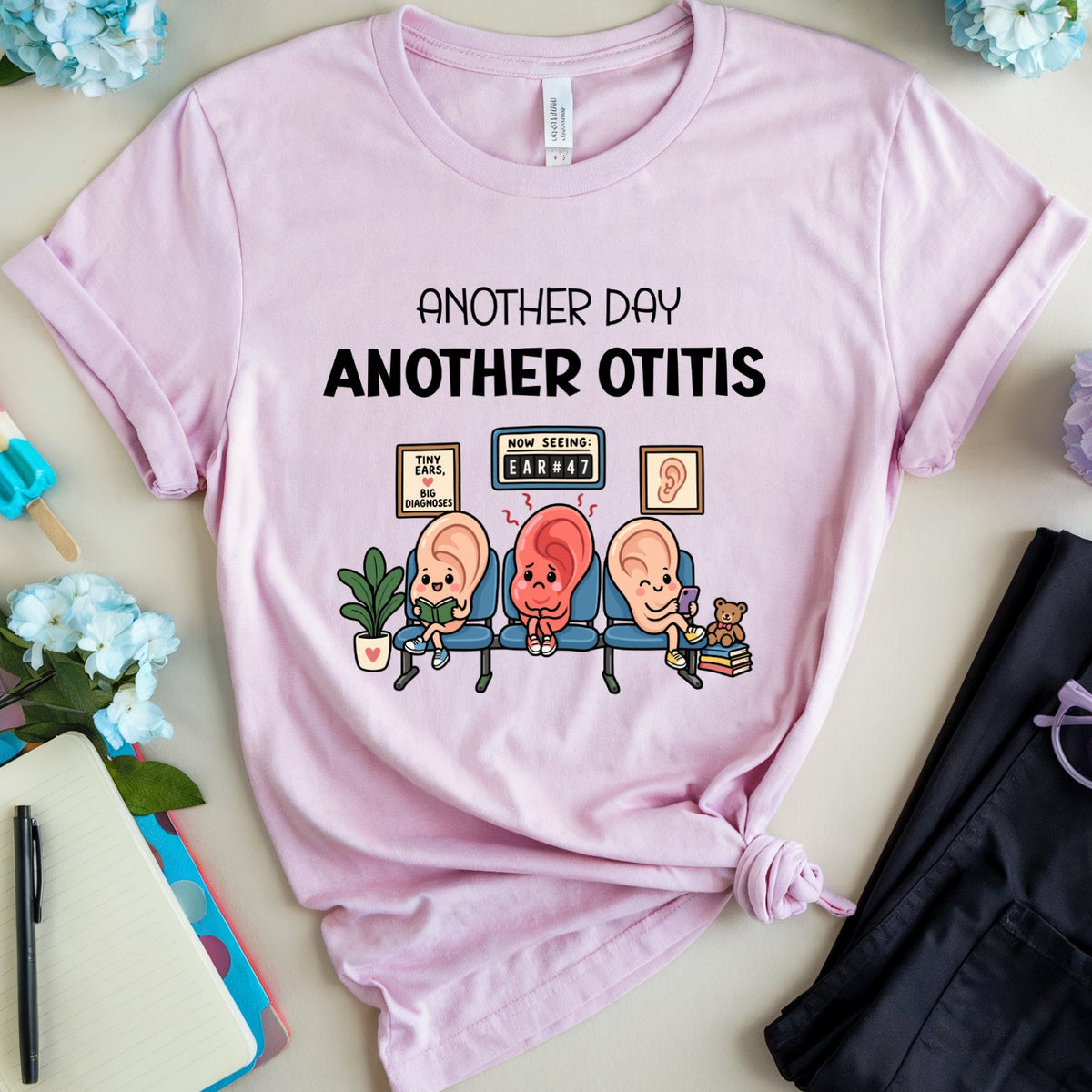 A light pink graphic t-shirt laid flat on a light beige surface, surrounded by decorative props including blue flowers, a notebook, a pen, a popsicle, and folded dark pants with purple glasses. The shirt features the text 'ANOTHER DAY ANOTHER OTIS' above a cartoon illustration of three otolaryngology doctors sitting on a couch.