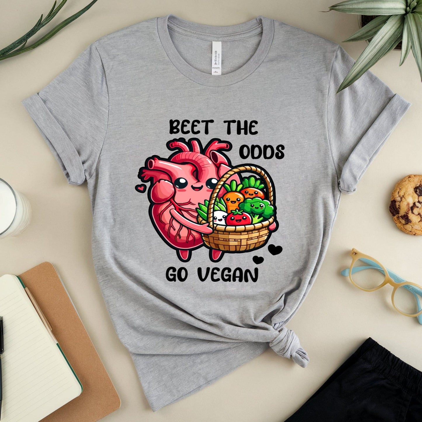 Gray t-shirt for cardiology with a heart and vegetables graphic and text 'Beet the Odds, Go Vegan' on a table with a glass of milk, cookies, and glasses.