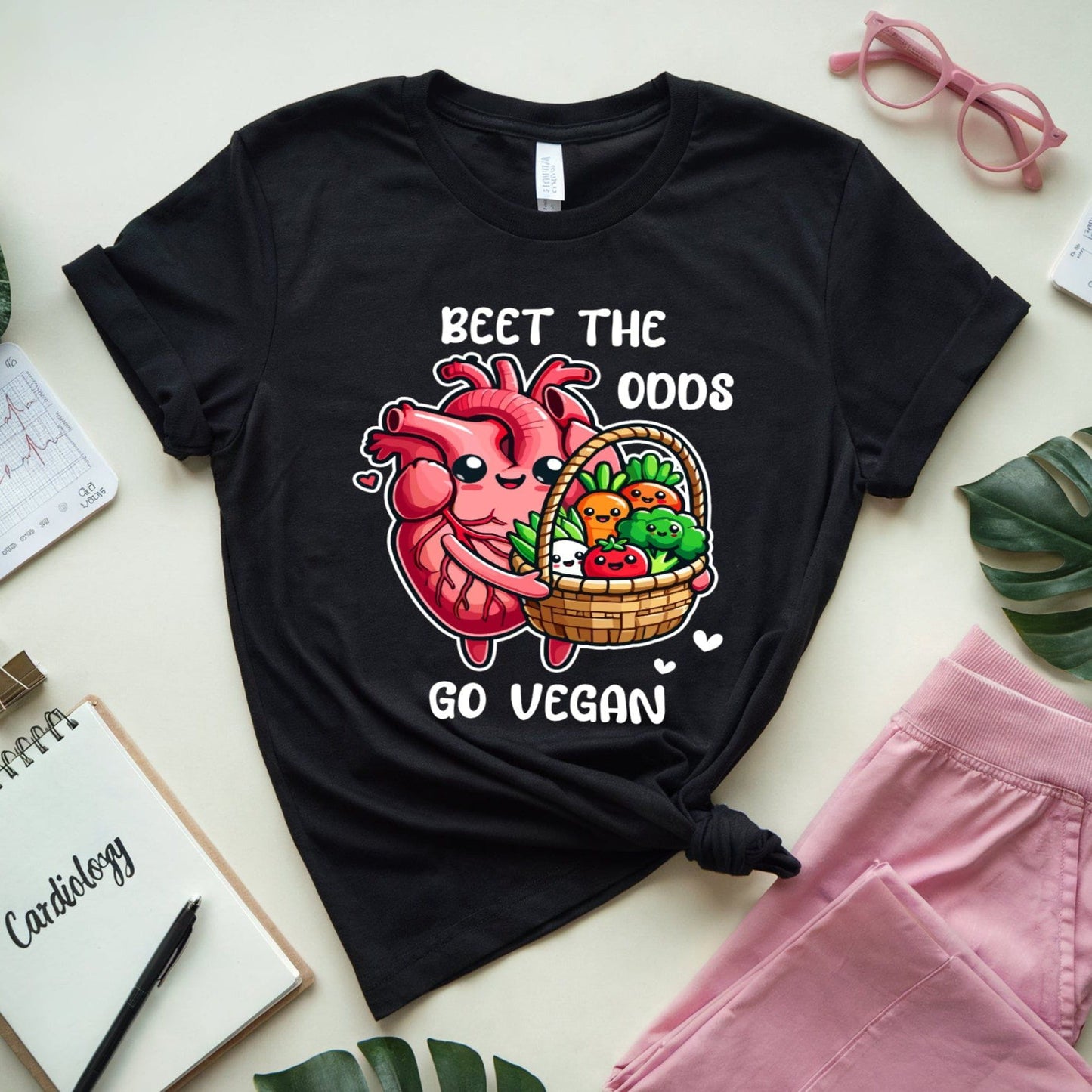 Black t-shirt for cardiology with a heart and vegetables graphic and text 'Beet the Odds, Go Vegan' on a light surface with pink pants and green leaves.