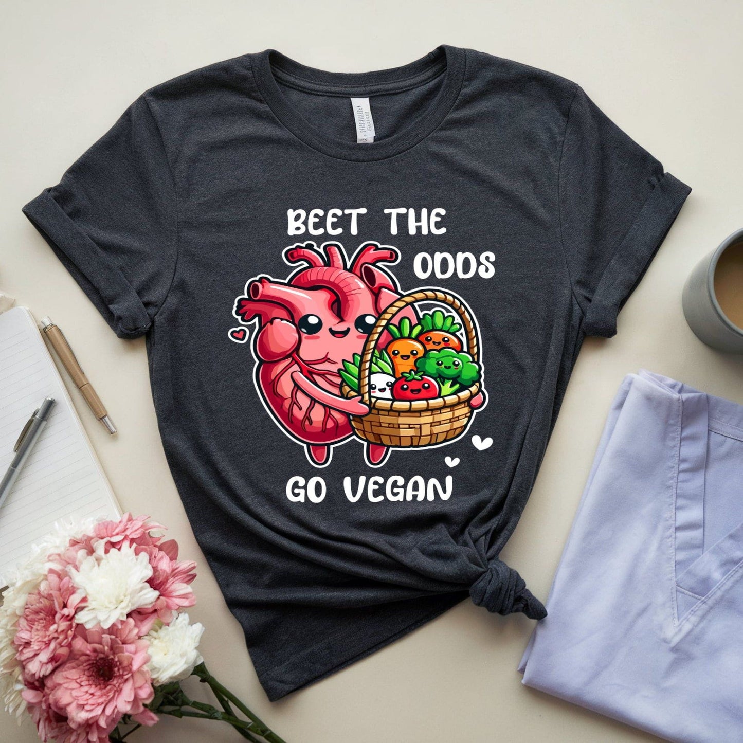 Dark heather gray t-shirt for cardiology with a heart and vegetables graphic, text 'Beet the Odds Go Vegan', placed on a light surface with flowers, jeans, and a mug.
