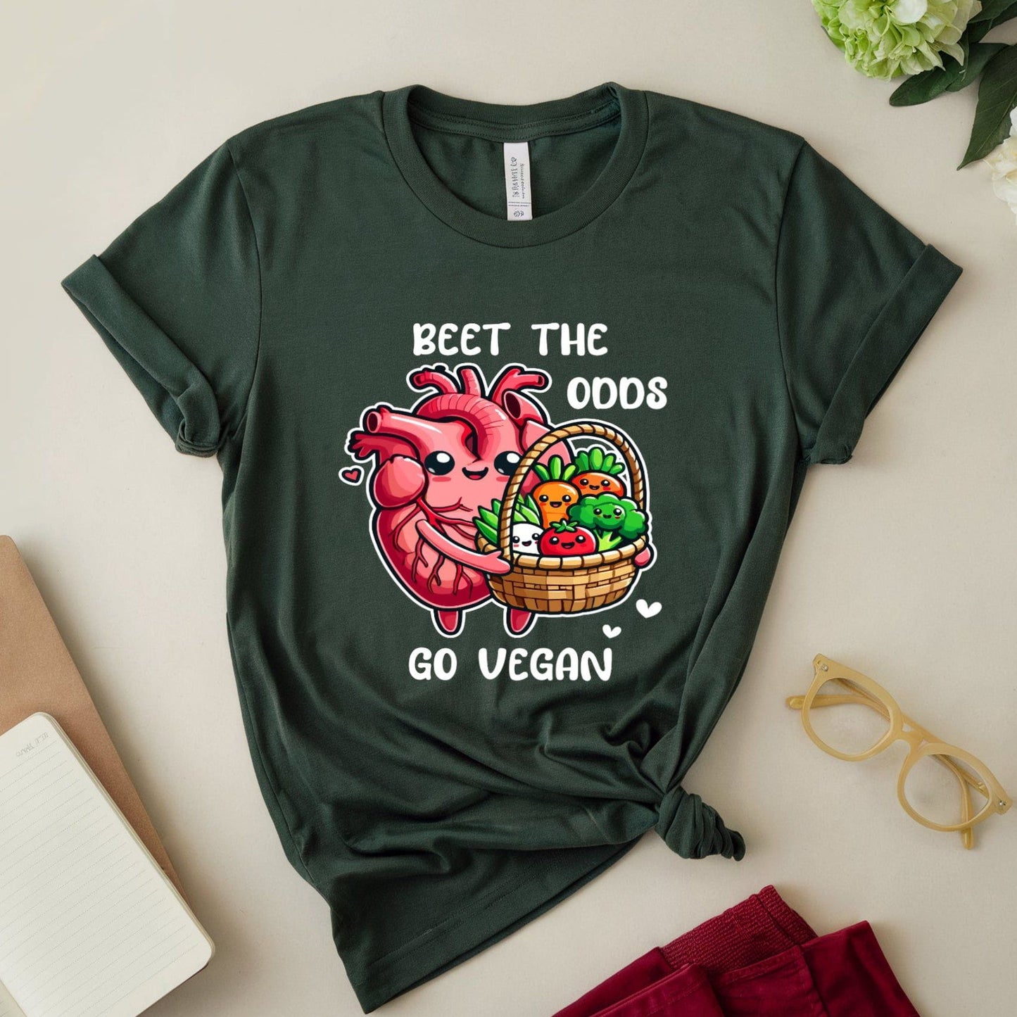 Forest green t-shirt for cardiology with a heart and vegetables graphic and text 'Beet the Odds, Go Vegan' on a light surface with flowers and books.
