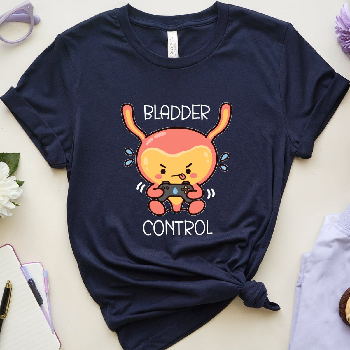 A navy blue graphic t-shirt with a cartoon bunny holding a phone and the words 'BLADDER CONTROL' is styled in a flat lay with purple flowers, glasses, a notebook, a drink, and folded light purple pants on a light beige background.