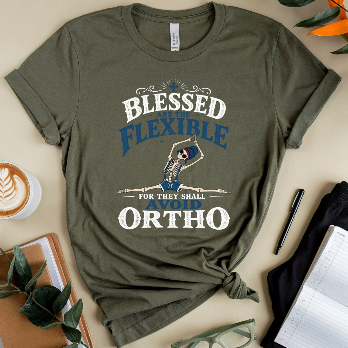 An olive green graphic t-shirt is presented in a styled flat-lay arrangement on a beige background. The t-shirt features a graphic of a person stretching with a bone staff, surrounded by the words 'BLESSED FLEXIBLE FOR THEY SHALL ORTHO' in white and blue.