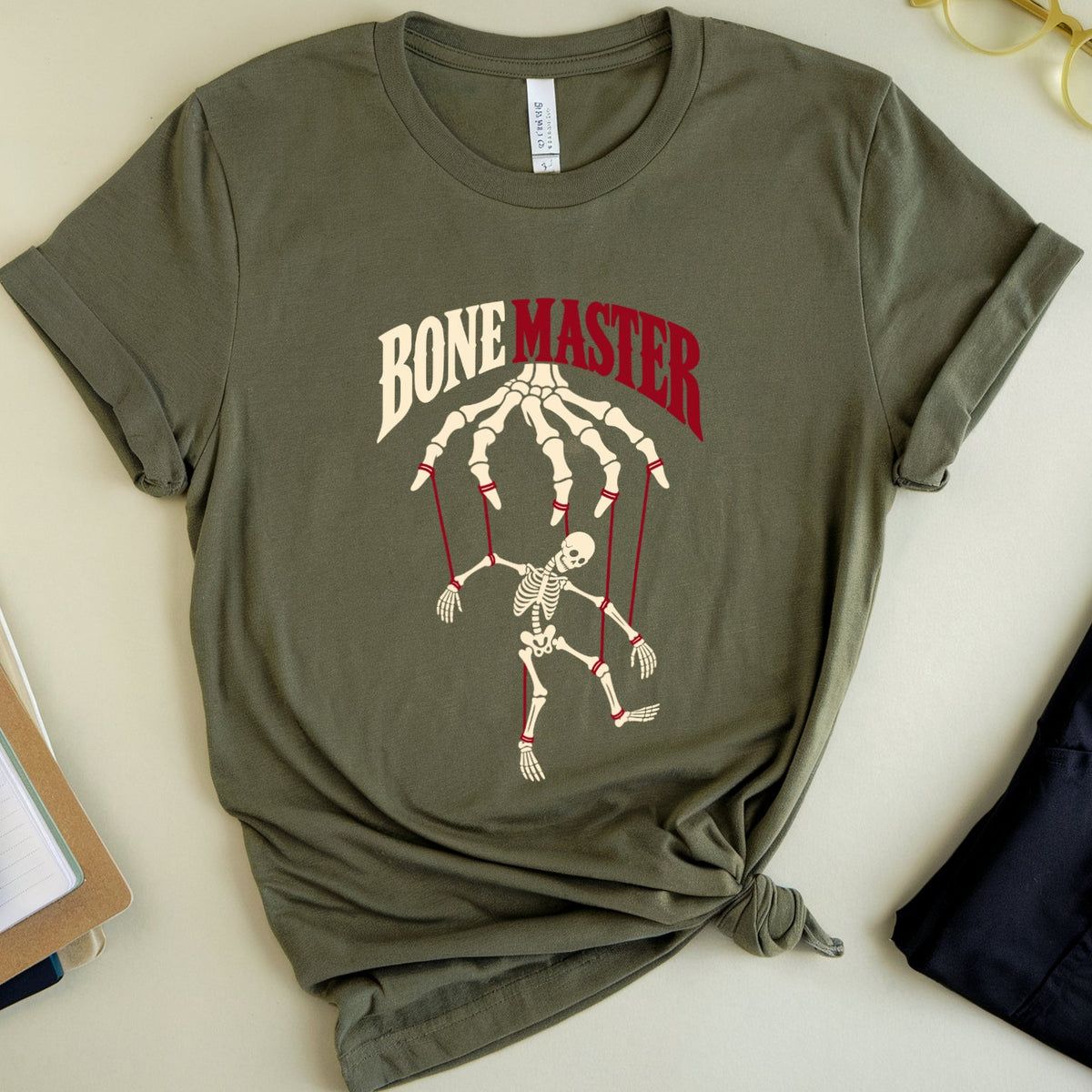 An olive green graphic t-shirt styled in a flat lay with a knot at the bottom, featuring a marionette skeleton graphic and the text 'BONE MASTER'. The shirt is surrounded by props including a pair of gold-rimmed glasses, a notebook, and a pair of dark pants on a light beige background.