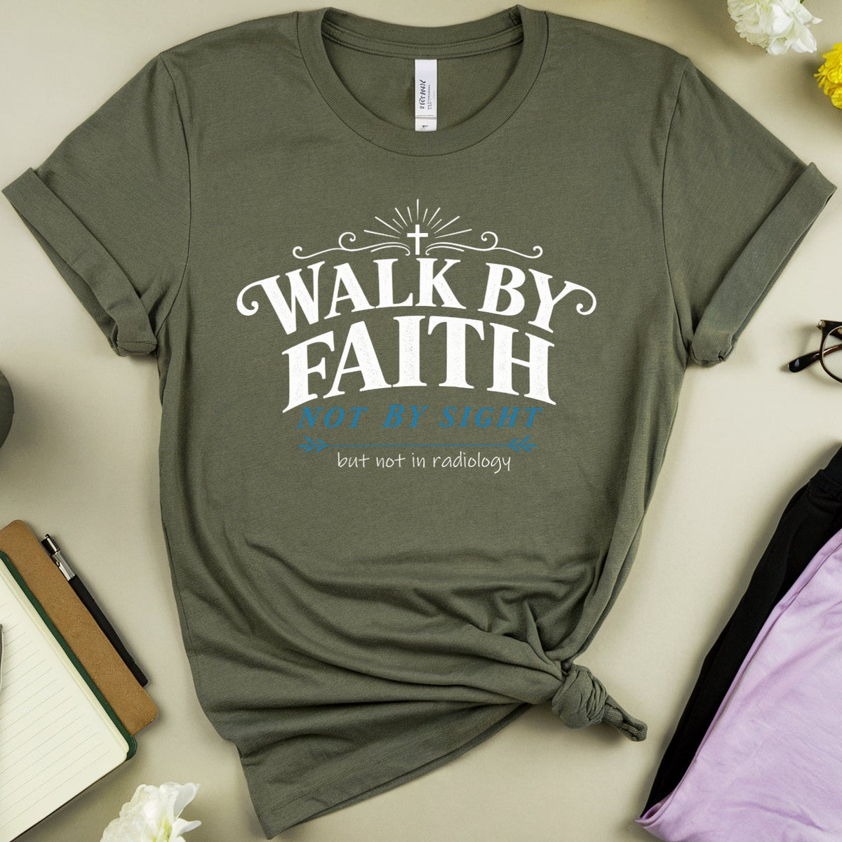 An olive green graphic t-shirt with a white and blue text print reading 'WALK BY FAITH get up strong but not in weakness', styled in a flat-lay arrangement with flowers, glasses, a notebook, and a cup on a beige background.