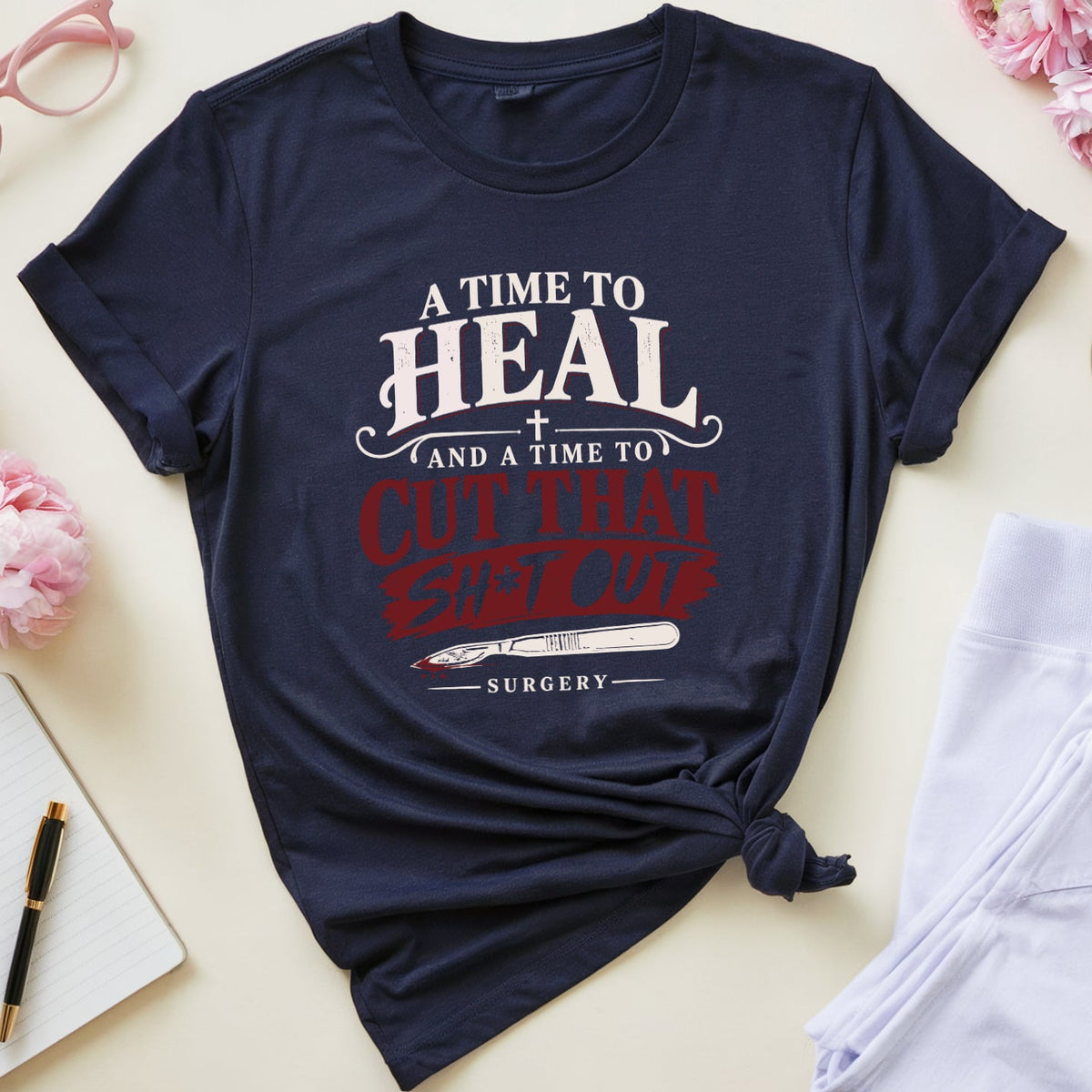 A navy blue graphic T-shirt laid flat on a light cream background, styled with pink flowers, folded white scrubs, pink eyeglasses, a notebook with a pen, and a small cup of tea. The shirt has a printed slogan in white and red text reading, 'A TIME TO HEAL AND A TIME TO CUT THAT STITCH UP SURGERY.