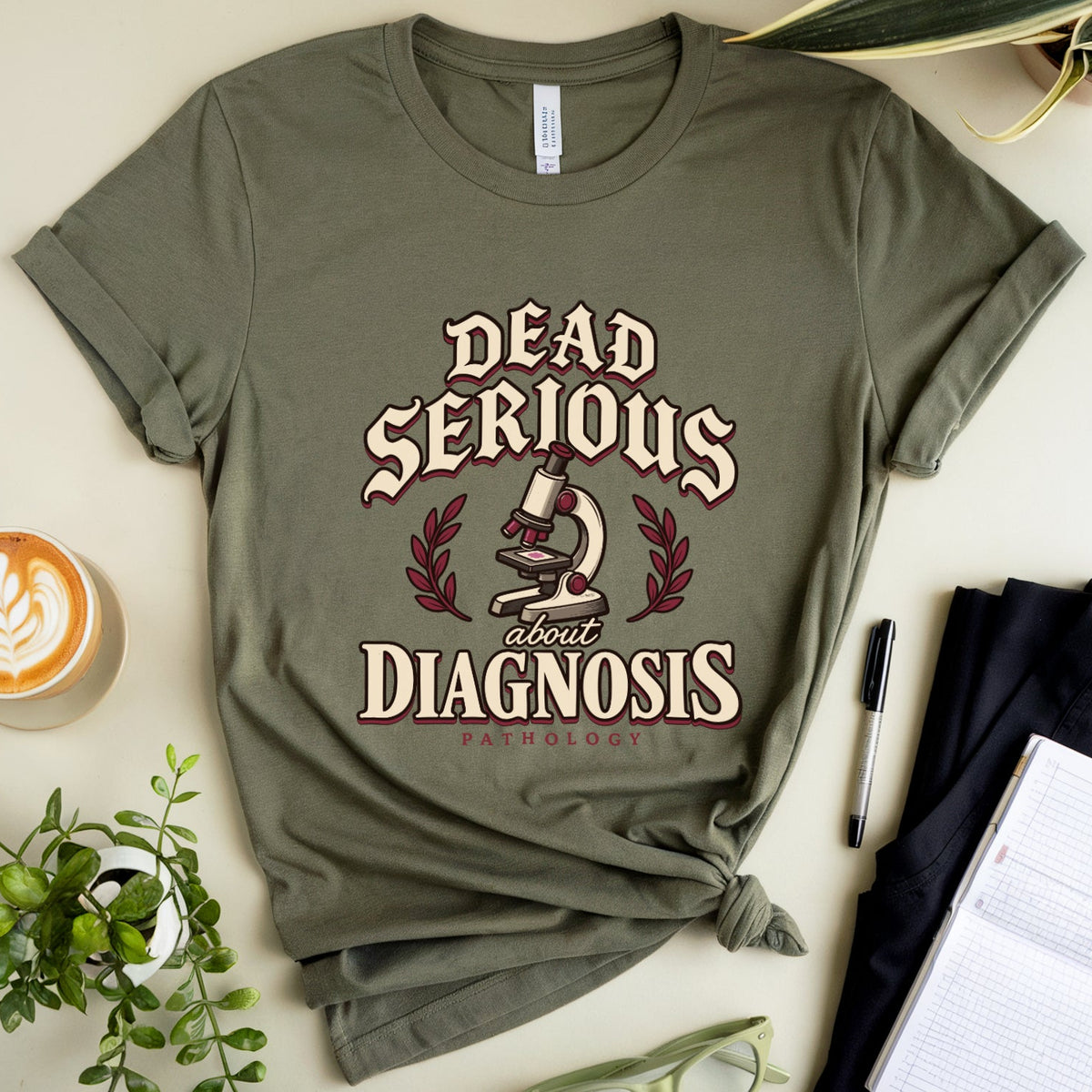 An olive green graphic t-shirt with the text 'DEAD SERIOUS about DIAGNOSIS' and a microscope illustration, styled in a flat lay with a coffee cup, notebook, glasses, pen, and plants on a light beige background.