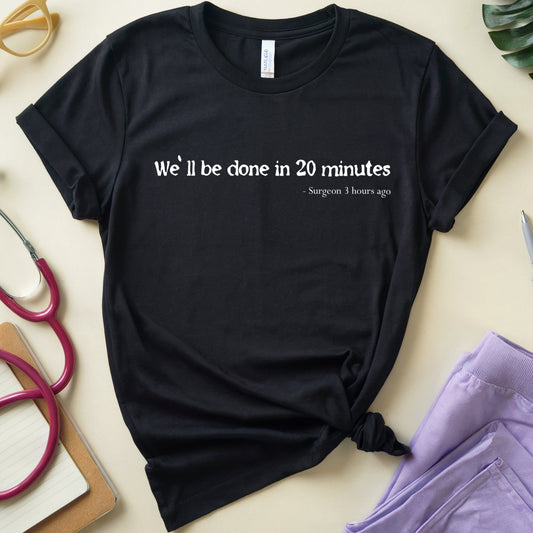 A black short-sleeve graphic T-shirt laid flat on a light beige surface, styled with props including yellow glasses, a stethoscope, a pen, a notebook, and folded lavender scrubs. The shirt has white text reading 'We'll be done in 20 minutes' with smaller text below, and the bottom hem is tied in a knot.