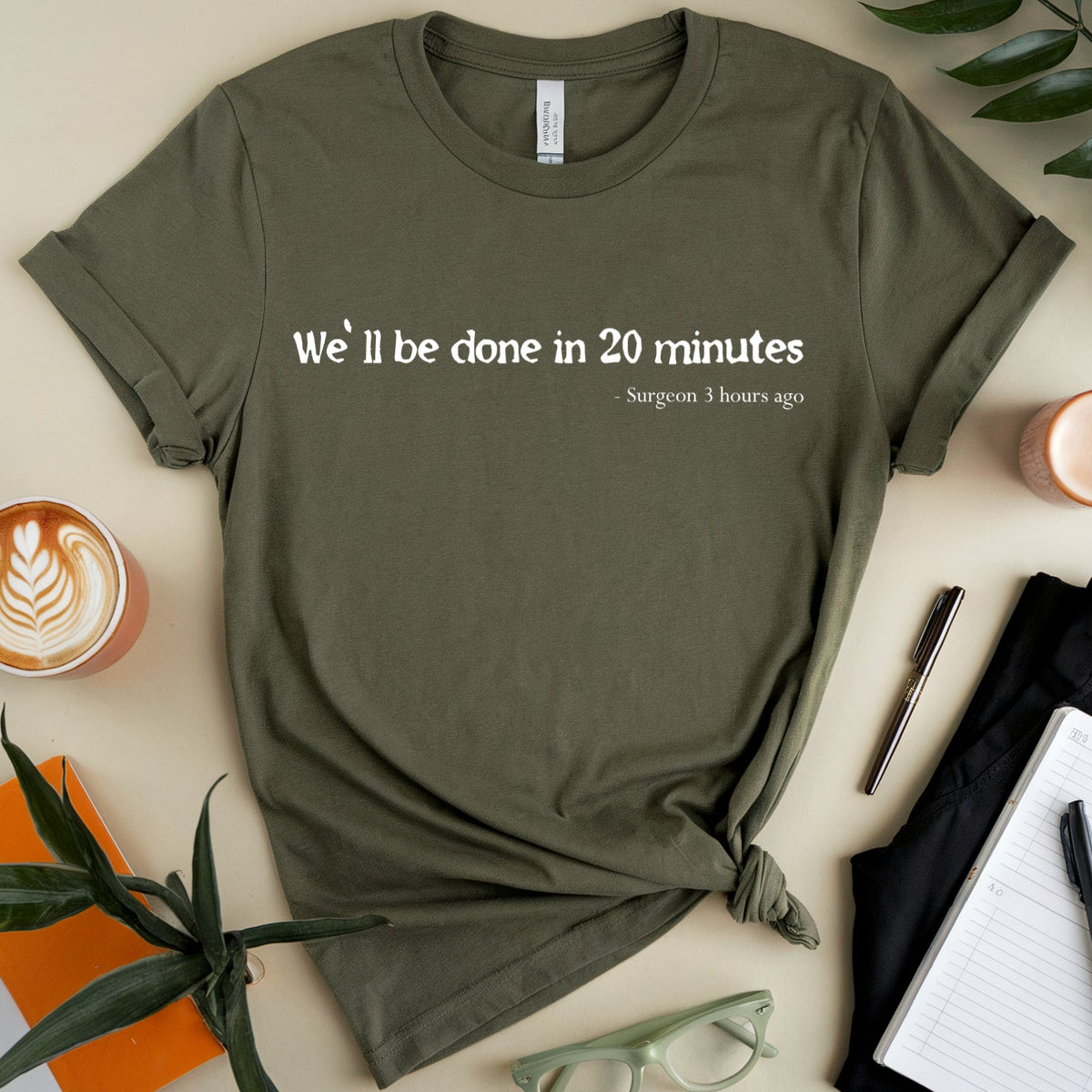 An olive green graphic t-shirt with the text 'We'll be done in 20 minutes' and 'S margram 3 hours ago' printed on the front, styled in a flat lay with a coffee cup, notebook, glasses, and plants on a beige background.