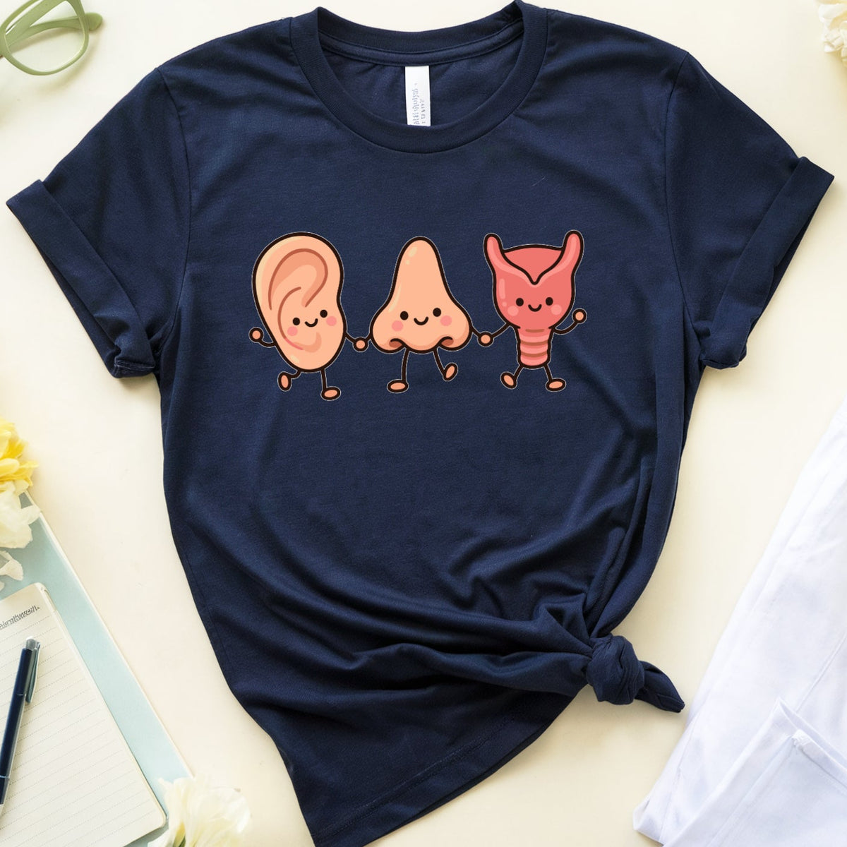 A navy blue graphic t-shirt featuring cartoon illustrations of an ear, a nose, and a throat in peach and orange, styled in a flat lay with yellow flowers, glasses, a notebook, a popsicle, and a folded white shirt on a light beige background.
