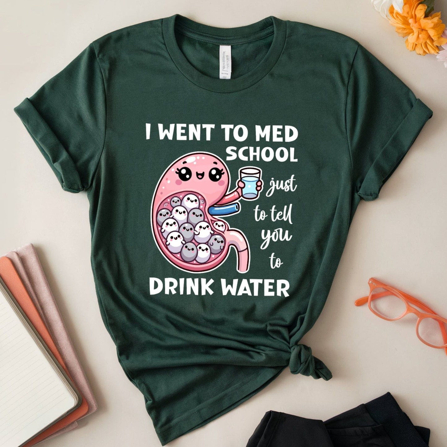 Green t-shirt with a humorous design about med school and water, placed on a light surface with flowers and glasses.