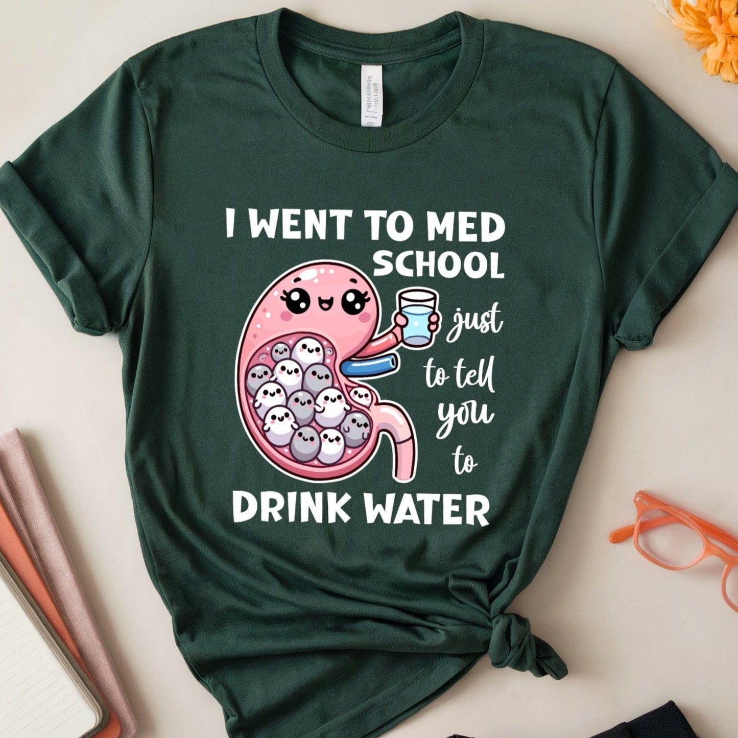 Green t-shirt with a humorous design about med school and water, placed on a light surface with flowers and glasses.