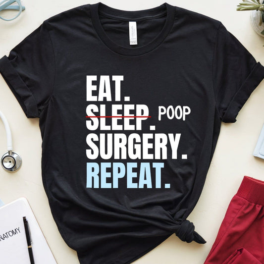 Black t-shirt with 'Eat. Sleep. Poop. Surgery. Repeat.' text on a light surface with medical-themed items.