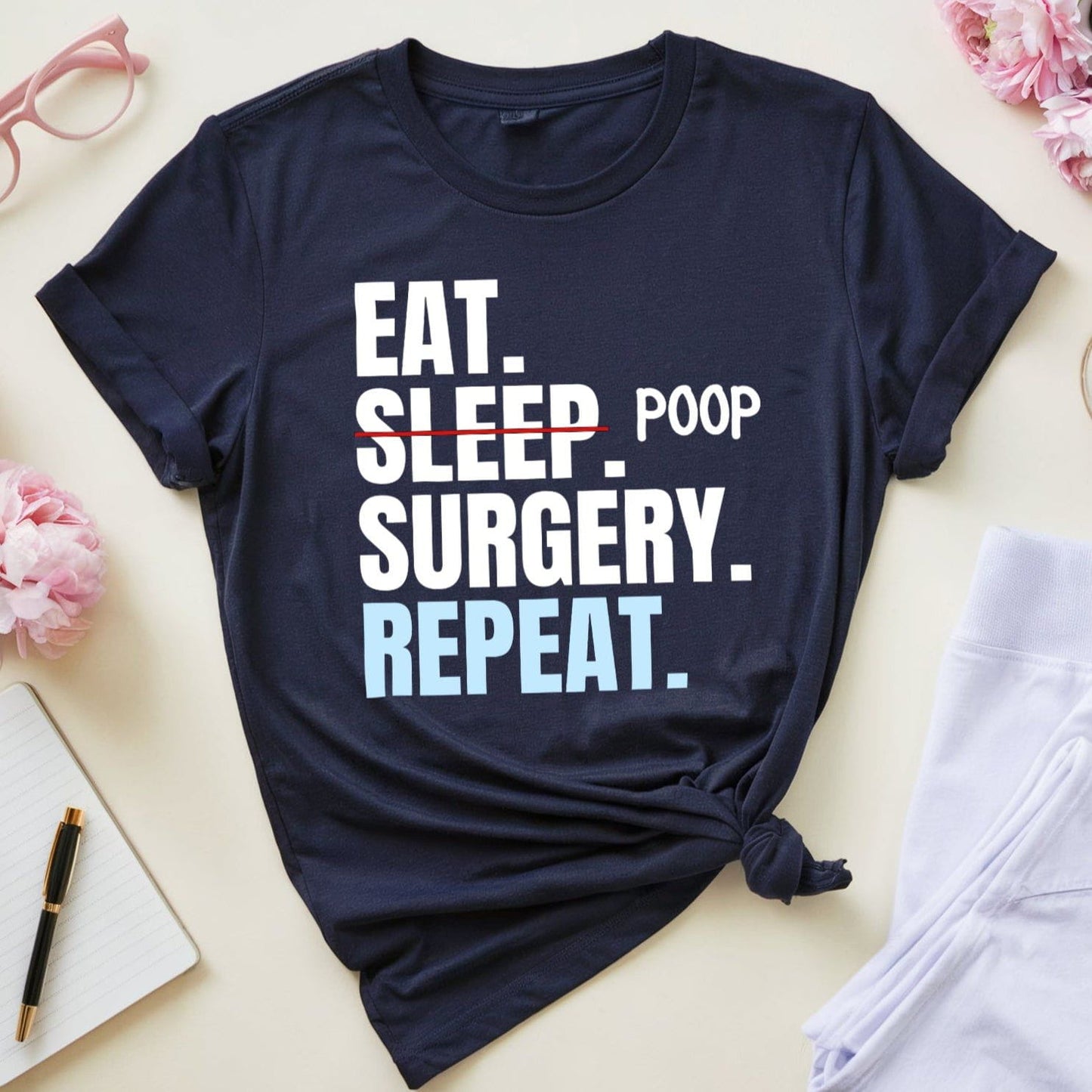 Navy blue t-shirt with 'Eat. Sleep. Poop. Surgery. Repeat.' text on a light surface with flowers and glasses.