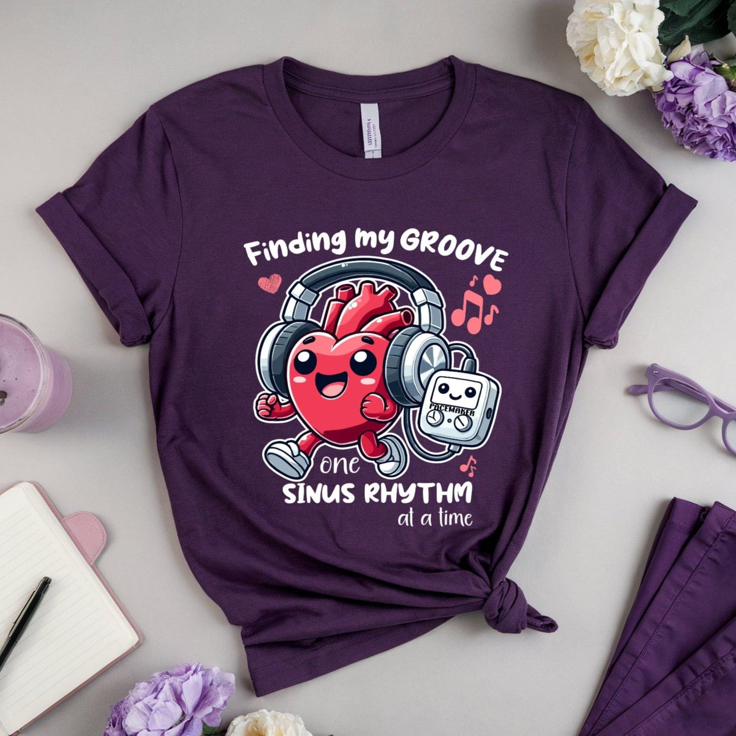 Purple t-shirt for cardiology with a jogging heart listening to music from its pacemaker design and text on a light gray surface with flowers and a drink.