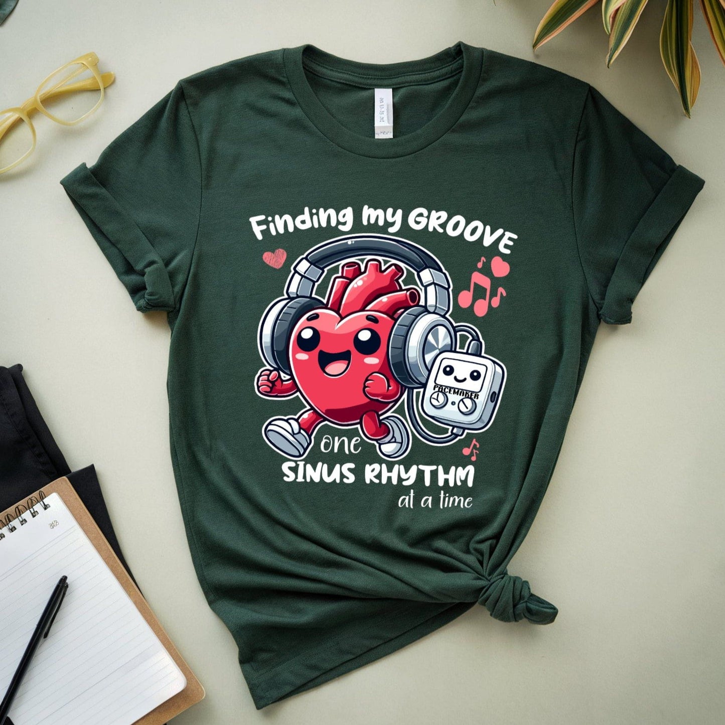 Green t-shirt for cardiology with a jogging heart listening to music from its pacemaker graphic and text on a light surface with a plant and notebook.
