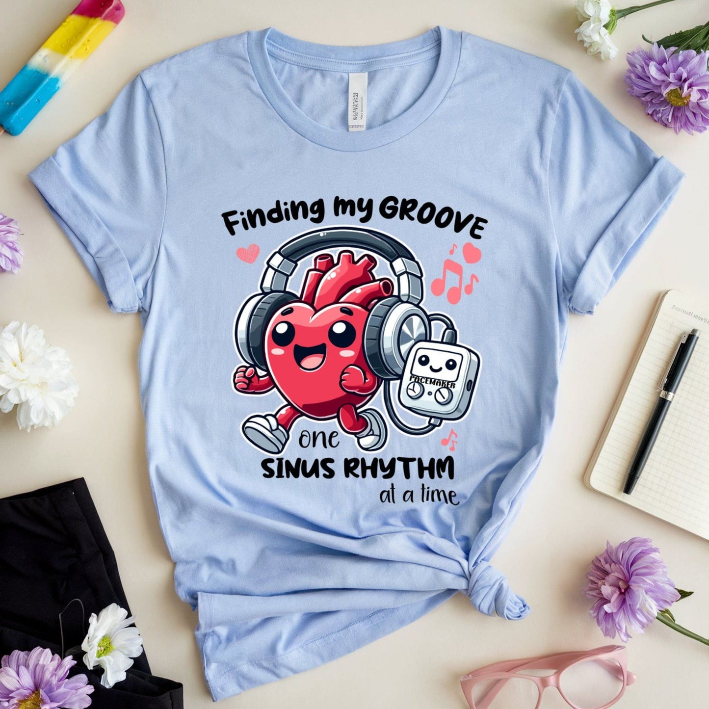 Light blue t-shirt for cardiology with a jogging heart listening to music from its pacemaker graphic and text on a light surface with flowers and sunglasses.