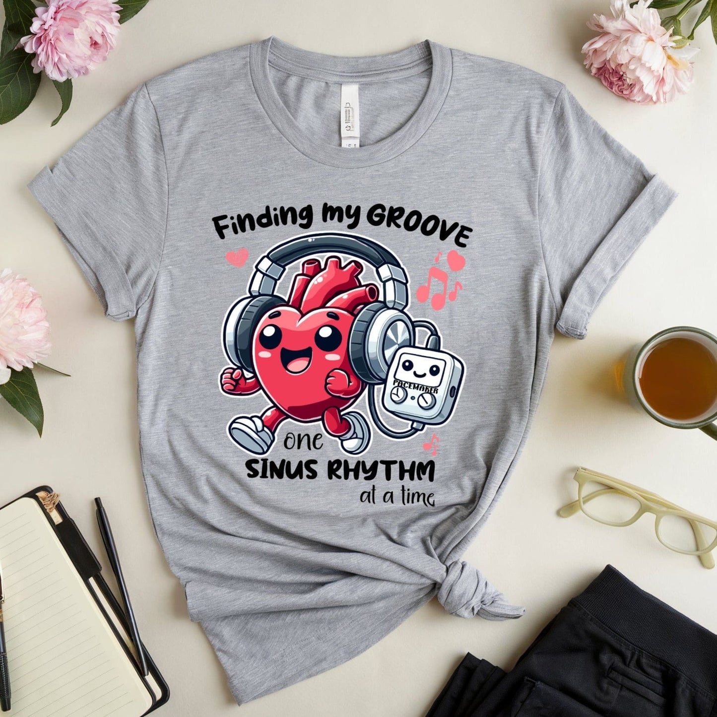 Gray t-shirt for cardiology with a jogging heart listening to music from its pacemaker design and text on a light surface with flowers and a cup of tea.