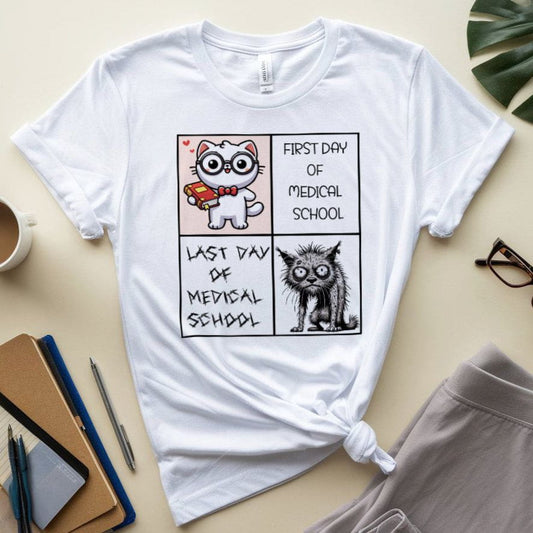 White t-shirt with a comic design of cats on the first and last day of medical school, placed on a surface with stationery items.