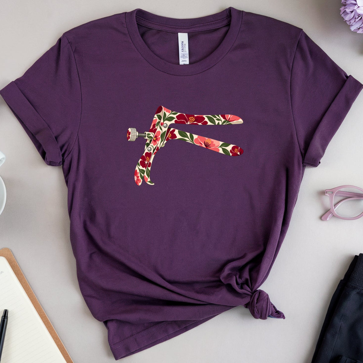 A flat-lay photograph of a dark purple short-sleeve t-shirt with a colorful floral graphic of a stylized figure on the chest. The shirt is styled in a flat-lay arrangement with a cup of latte, a notebook, pink eyeglasses, and purple and white flowers on a light gray background.