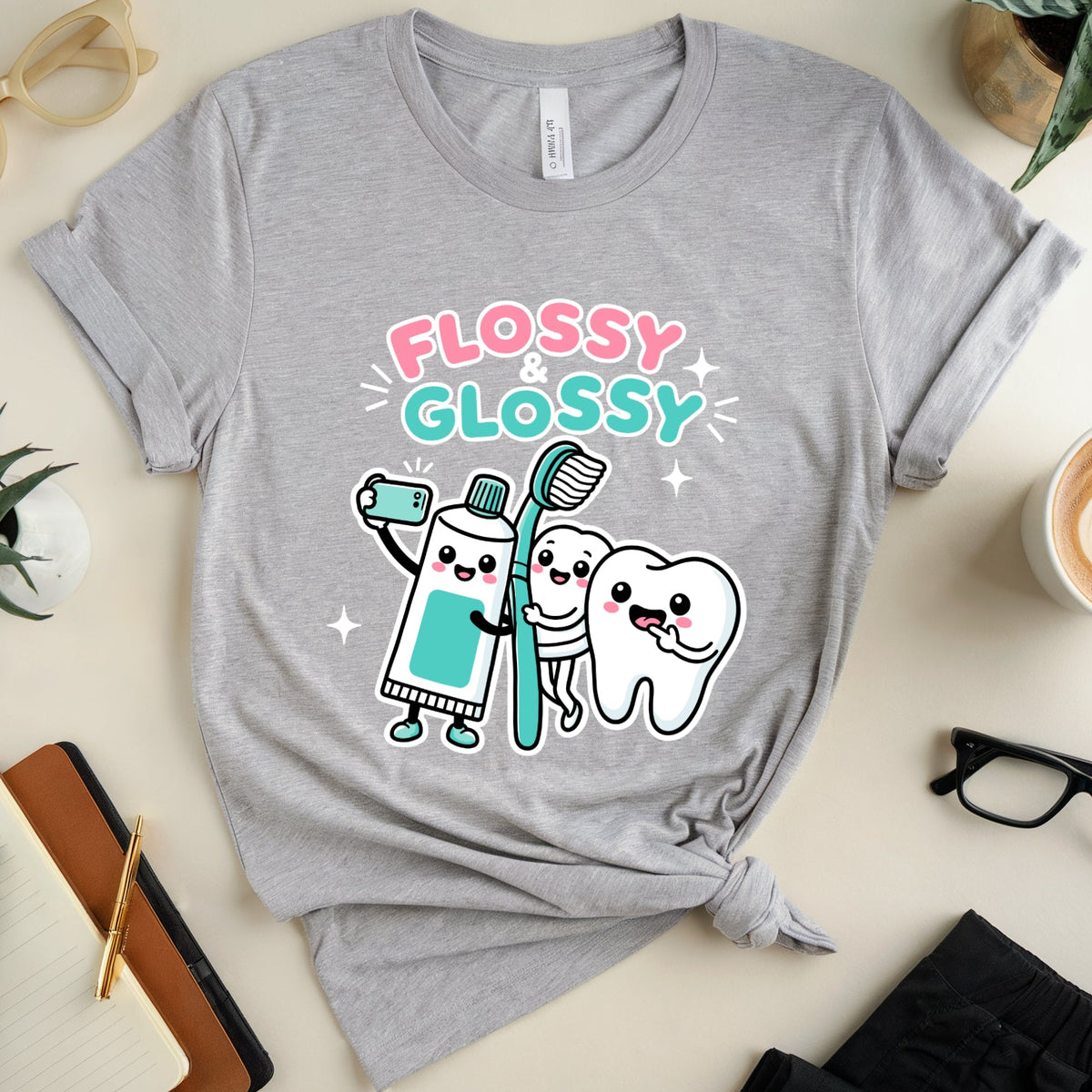 A heather gray graphic t-shirt featuring a cute cartoon design of a toothpaste tube and a tooth brushing teeth, with the text 'FLOSSY GLOSSY' in pink and teal. The t-shirt is styled in a flat lay with glasses, plants, a coffee cup, and notebooks on a light beige background.