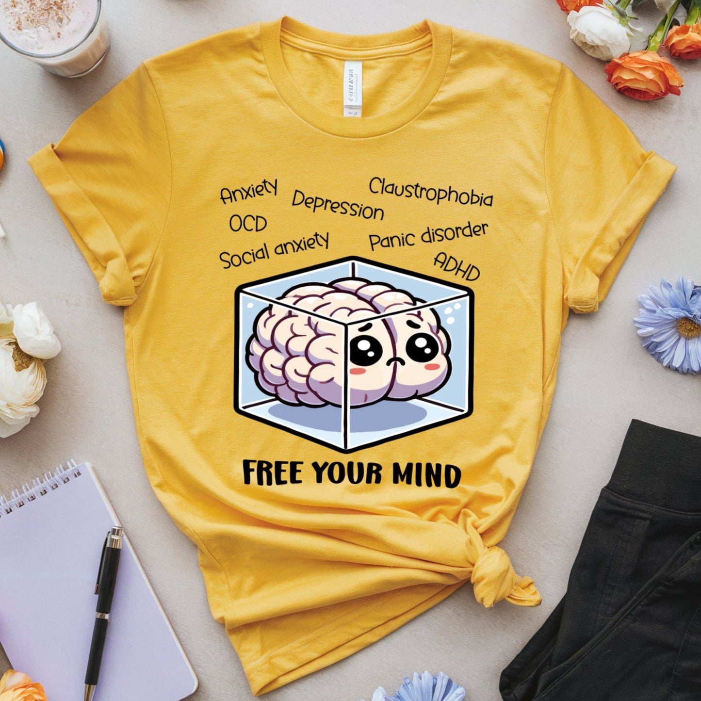 Yellow t-shirt with a brain graphic and text on a table with flowers, a notebook, and a drink.