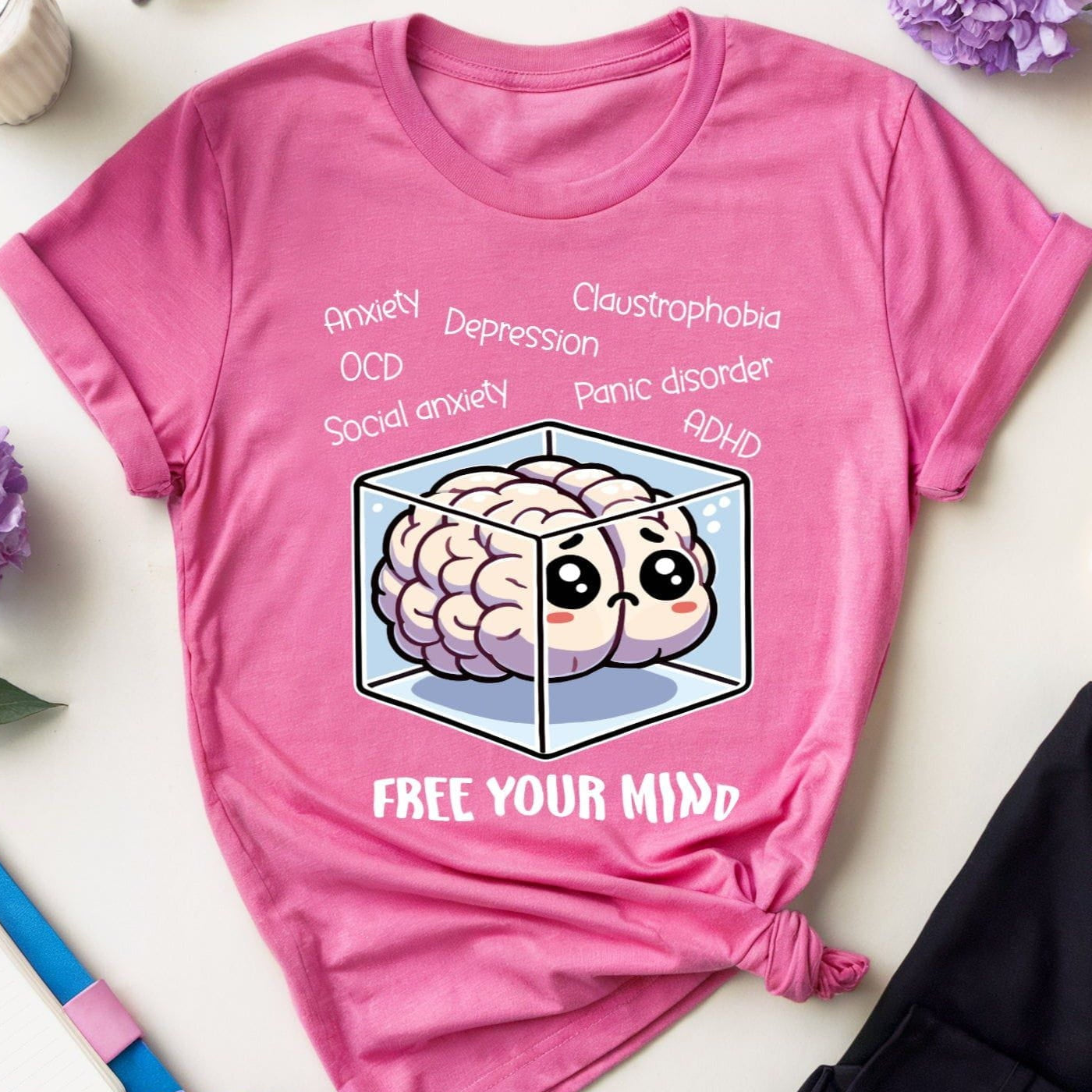 Pink t-shirt with brain graphic and text on a white surface with flowers and glasses.