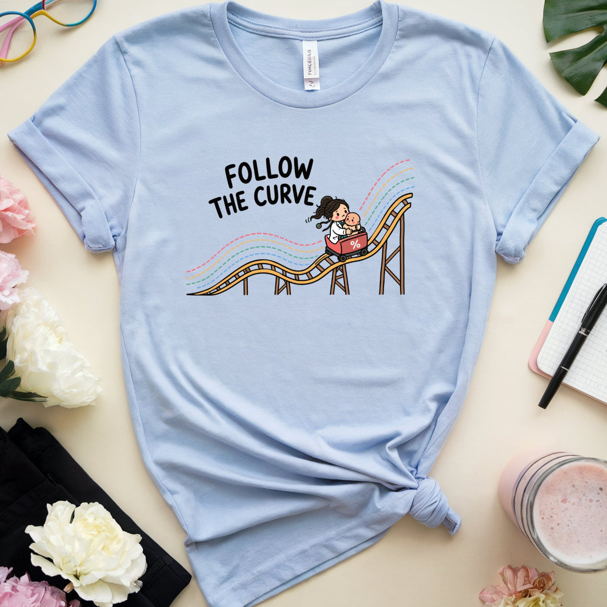A light blue graphic t-shirt featuring a cartoon roller coaster design with the text 'FOLLOW THE CURVE'. The t-shirt is styled in a flat-lay arrangement on a beige background with pink and white flowers, a pair of colorful sunglasses, a notebook, a drink, and a green leaf.