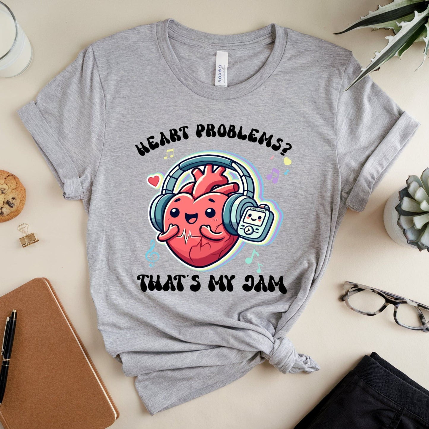 Gray t-shirt with a heart graphic and text on a table with a notebook, cookie, and glasses.