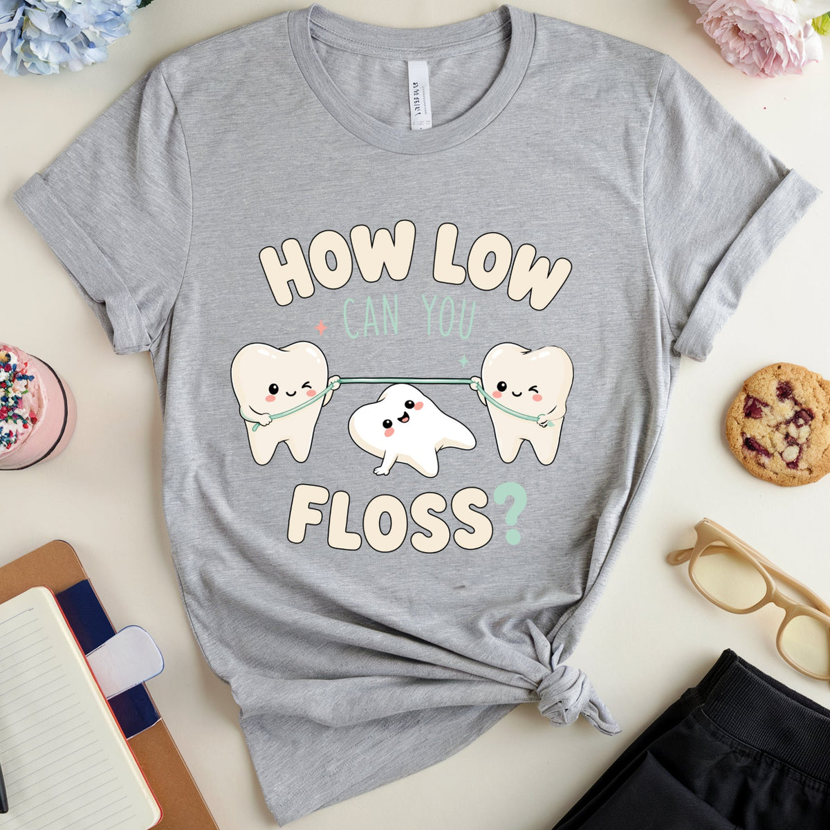 A heather gray graphic t-shirt with a cute cartoon tooth design and the text 'HOW LOW CAN YOU FLOSS?' styled in a flat-lay arrangement with flowers, glasses, a notebook, and a cookie on a light beige background.