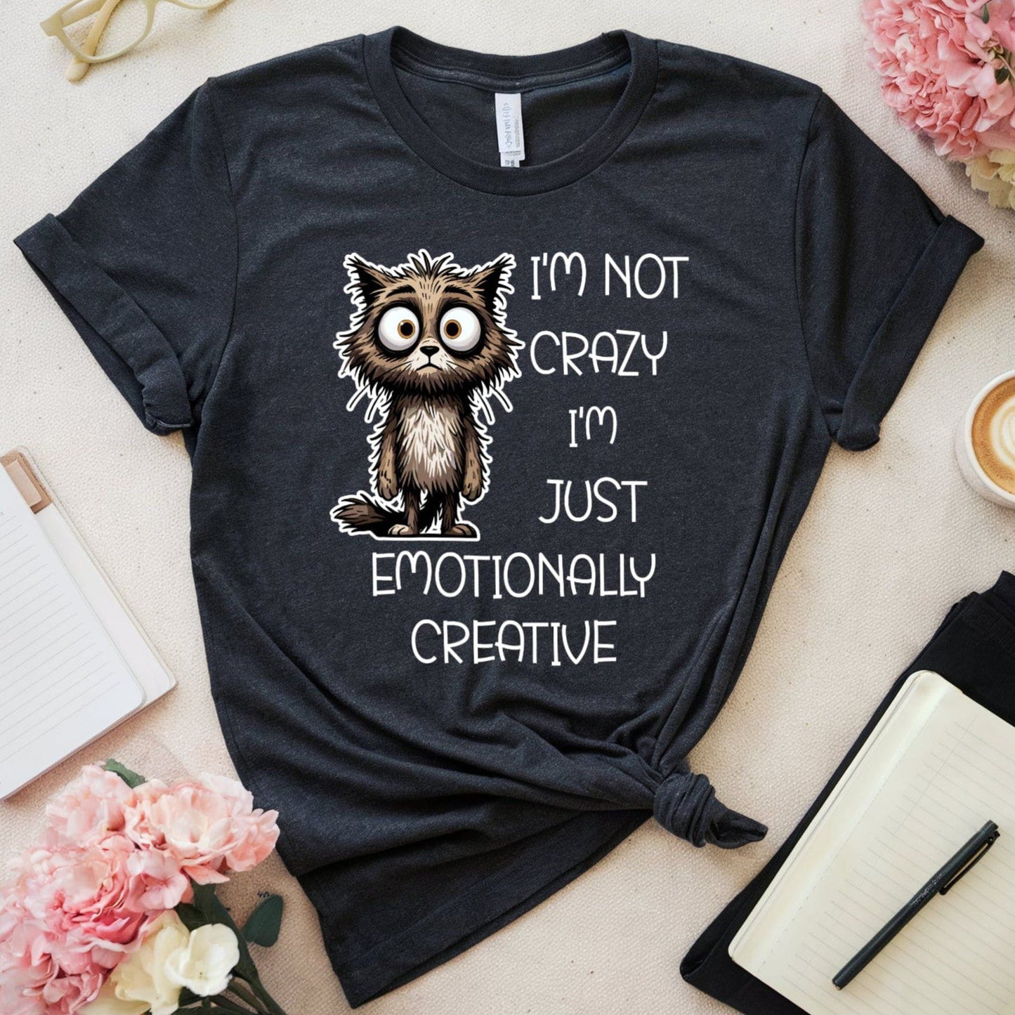 Black t-shirt with a cat graphic for mental health that is sarcastic on a light surface with flowers and a cup of coffee.