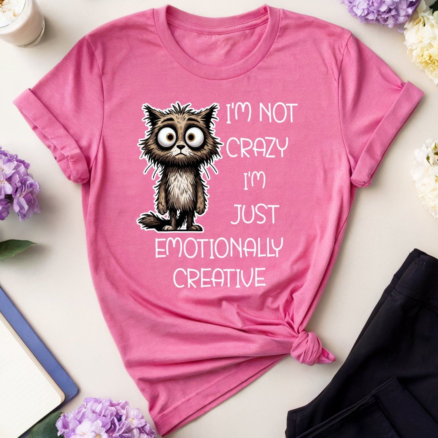 Pink t-shirt with a cat graphic and text on a table with flowers and glasses.