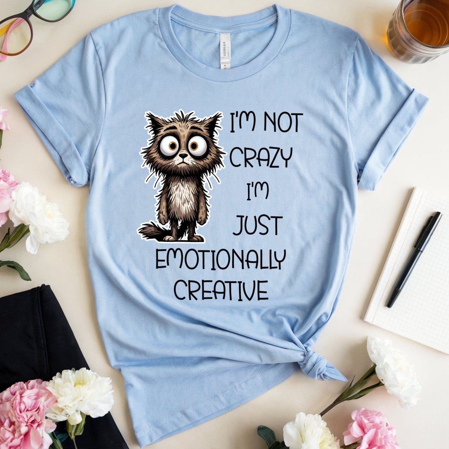 Light blue t-shirt with a cat graphic for mental health that is sarcastic on a table with flowers and glasses.