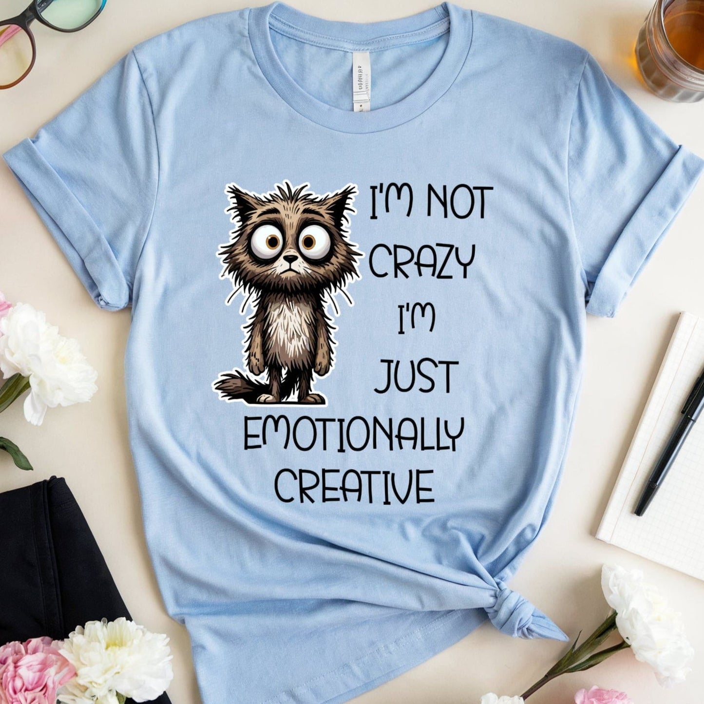 Light blue t-shirt with a cat graphic for mental health that is sarcastic on a table with flowers and glasses.