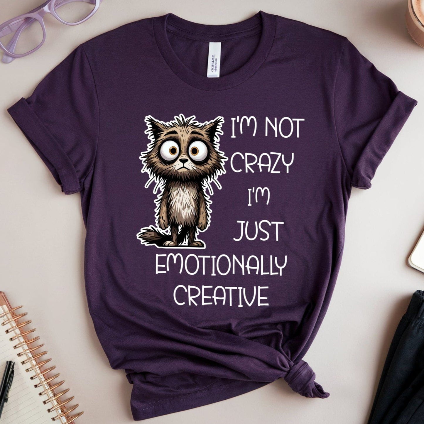 Purple t-shirt with a cat graphic for mental health that is sarcastic on a table with accessories