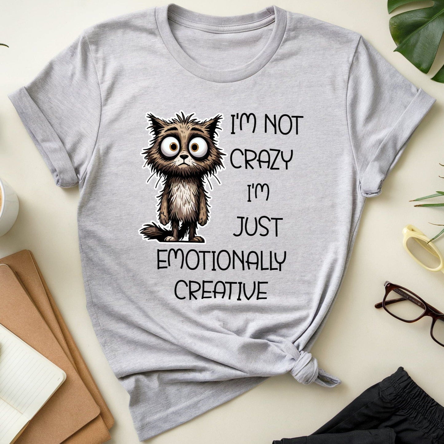 Gray t-shirt with a cat graphic for mental health that is sarcastic on a desk with a cup of coffee, notebook, and glasses.