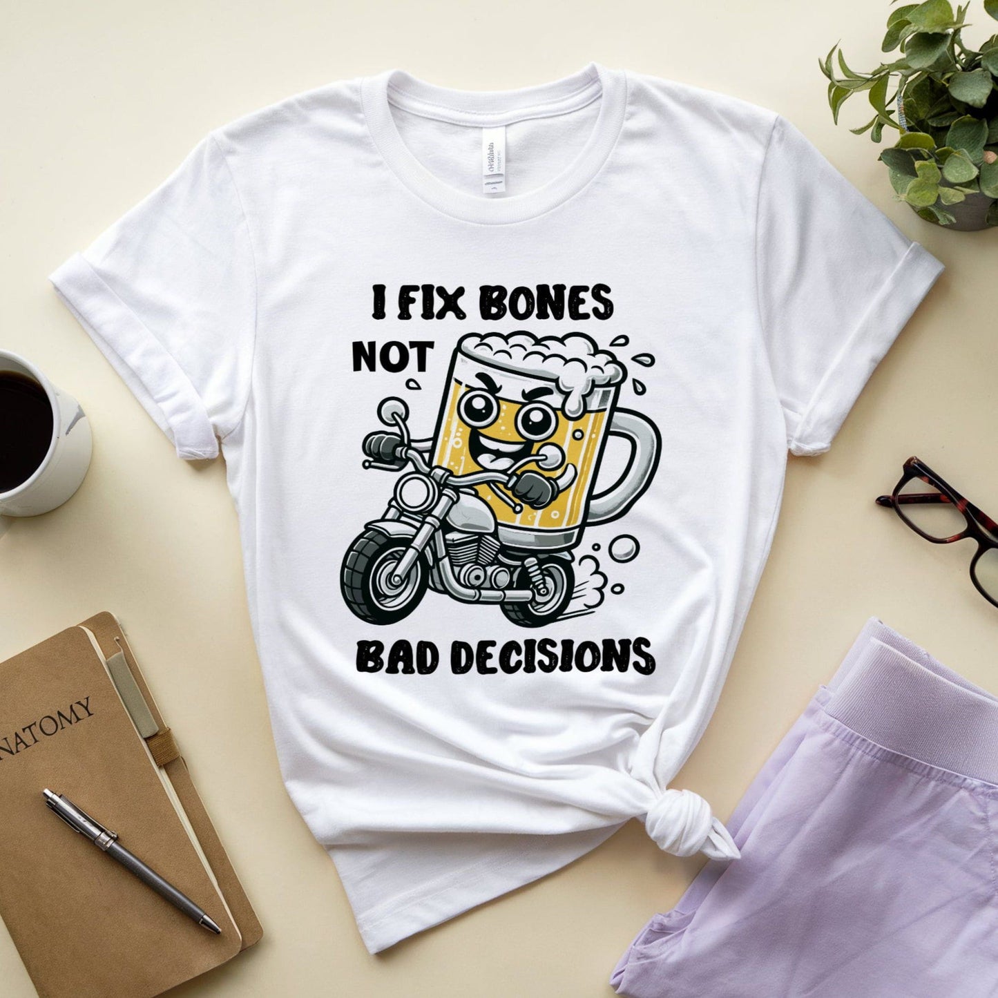 White t-shirt for orthopedic residents with a graphic design on a beige surface with a notebook, coffee cup, and glasses.