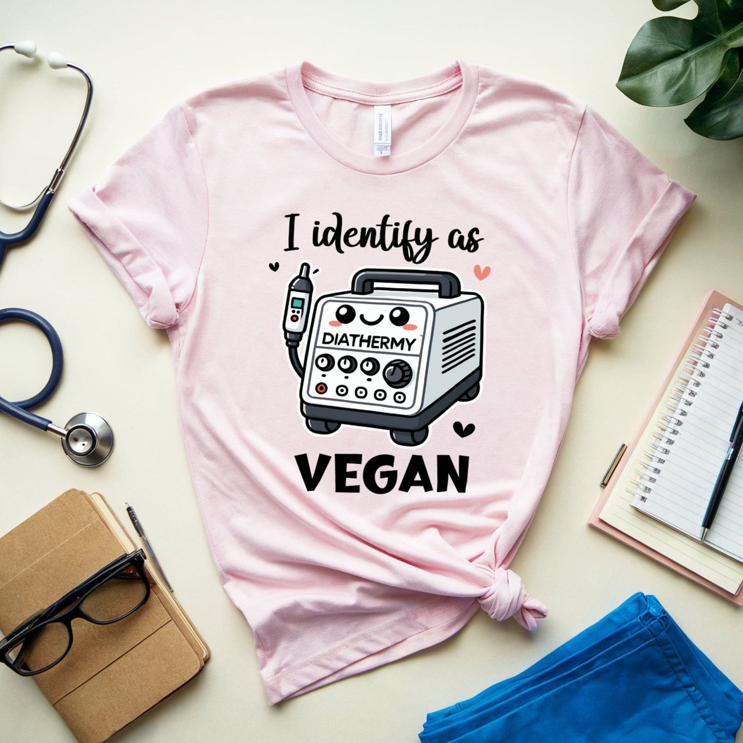 light pink t-shirt with a graphic and text 'I identify as DIATHERMY VEGAN' on a light surface with stationery items.