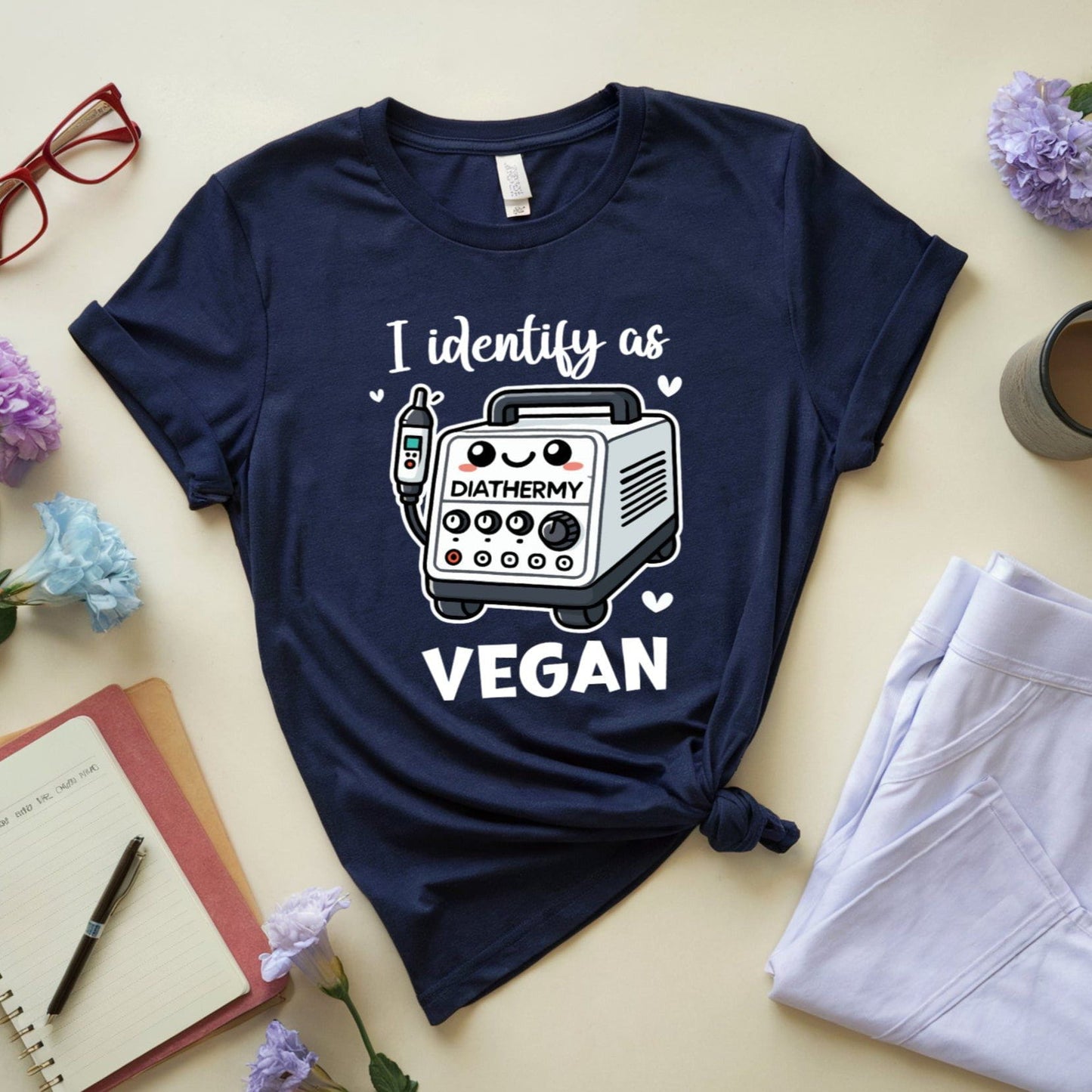 Navy blue t-shirt with 'I identify as vegan' graphic on a light surface with flowers, coffee, and pants.
