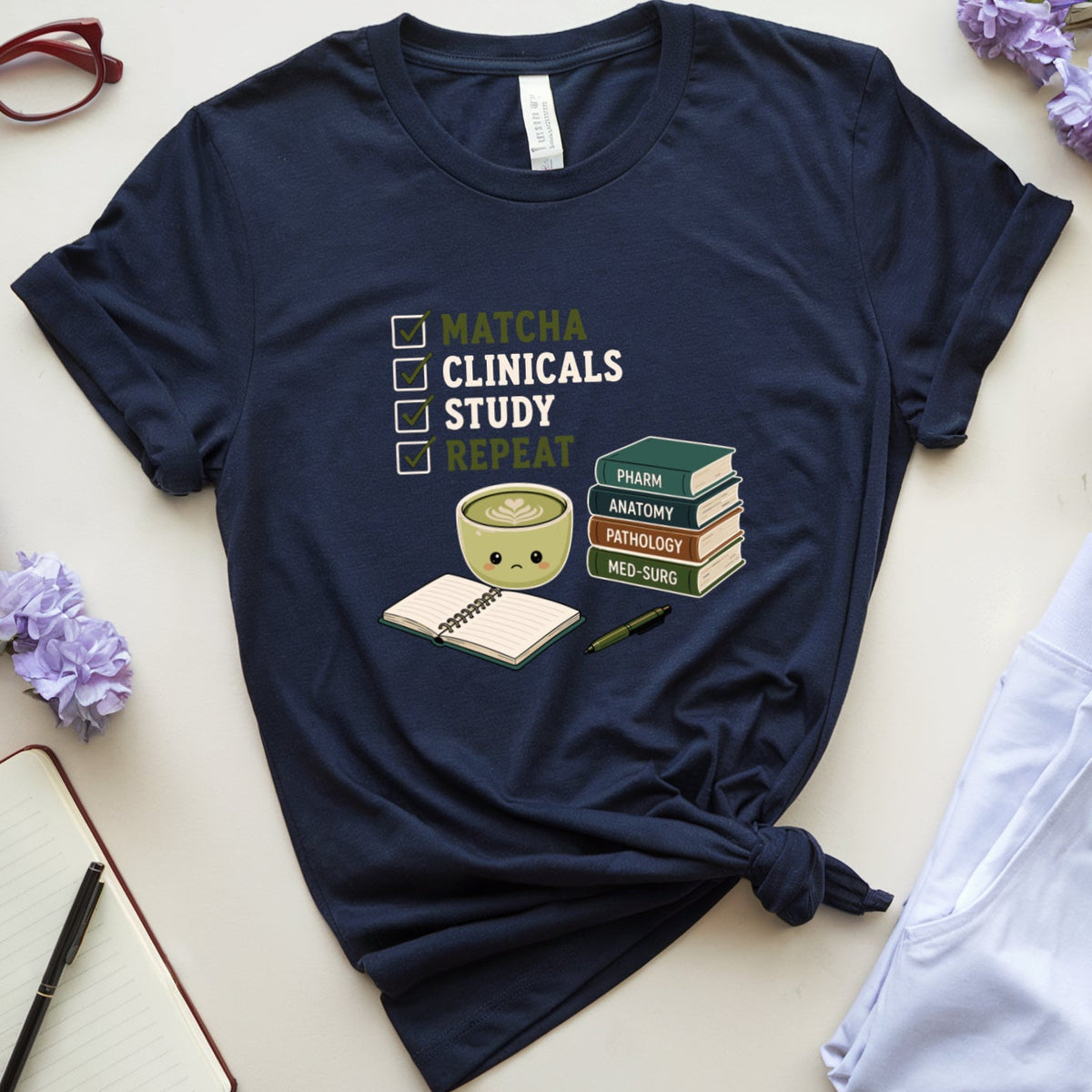 A navy blue graphic t-shirt styled in a flat lay with lavender flowers, glasses, a notebook, a cup of coffee, and white sweatpants on a light beige background. The t-shirt features a cute matcha bowl illustration and the text 'MATCHA CLINICALS STUDY REPEAT'.