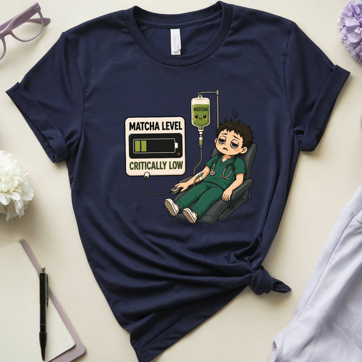 A navy blue graphic t-shirt laid flat on a light beige surface, styled with glasses, flowers, a notebook, a pen, a mug of coffee, and light gray sweatpants. The t-shirt features a cartoon graphic of a tired nurse with a low battery and an IV bag labeled 'MATCHA LEVEL CRITICALLY LOW'.