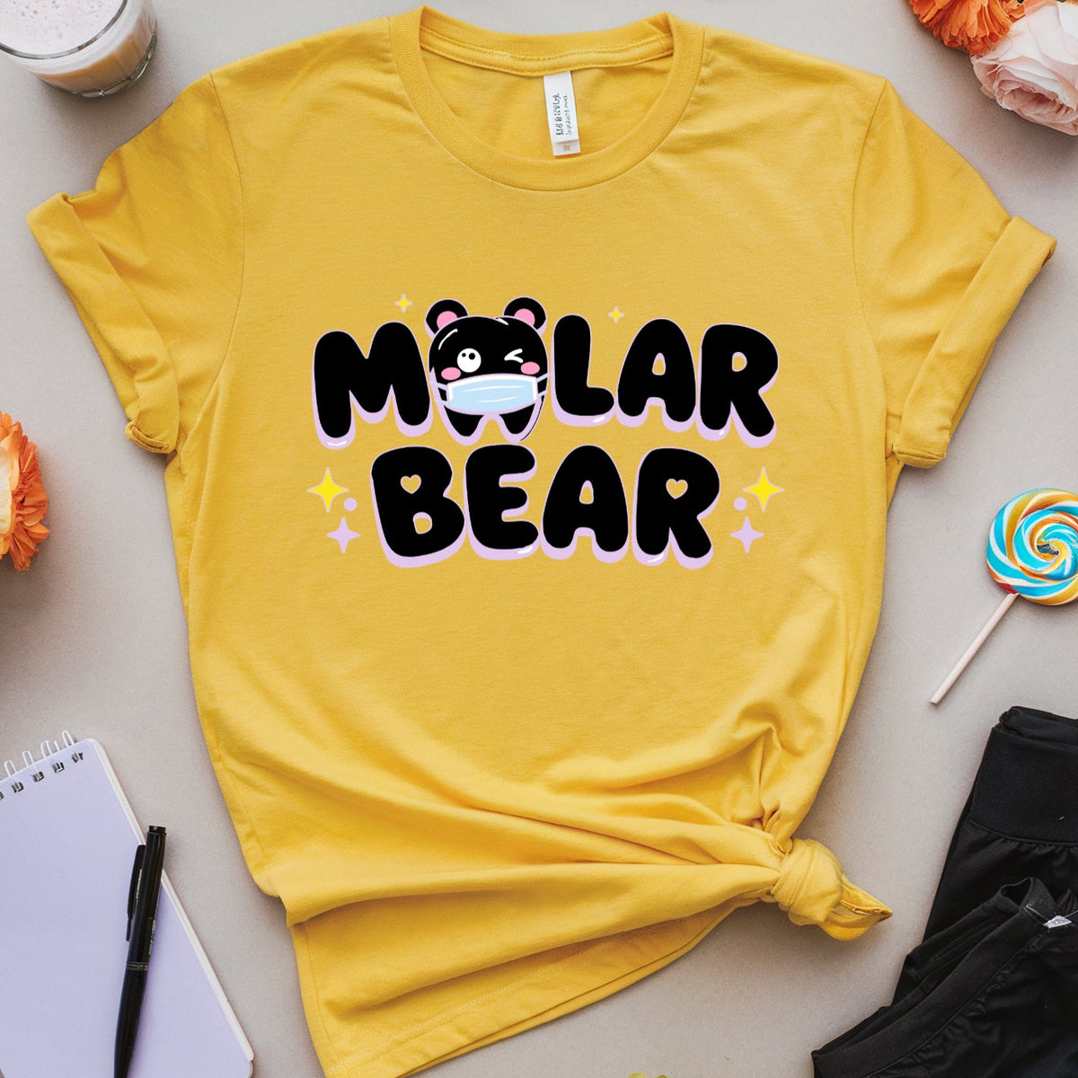 A yellow graphic T-shirt laid flat on a beige surface, surrounded by flowers, sunglasses, a drink, a notebook, a pen, a lollipop, and black shorts. The shirt has a large black 'Molar Bear' text graphic with a bear face and small star accents.