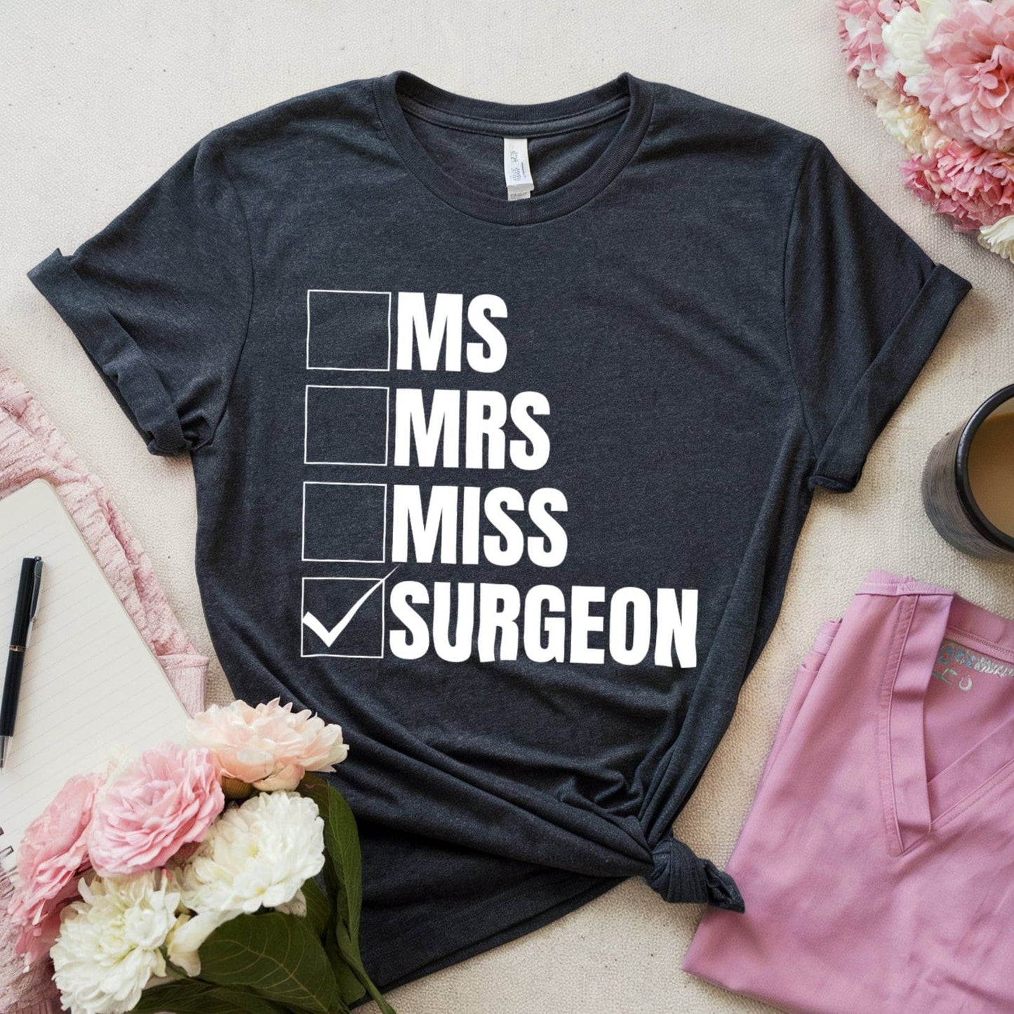 Dark heather grey t-shirt with 'MS Mrs Miss Surgeon' text, pink shorts, coffee cup, and flowers on a light surface