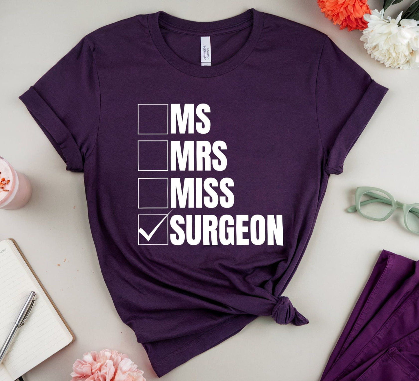 Purple t-shirt with humorous text design on a light surface with flowers and glasses.